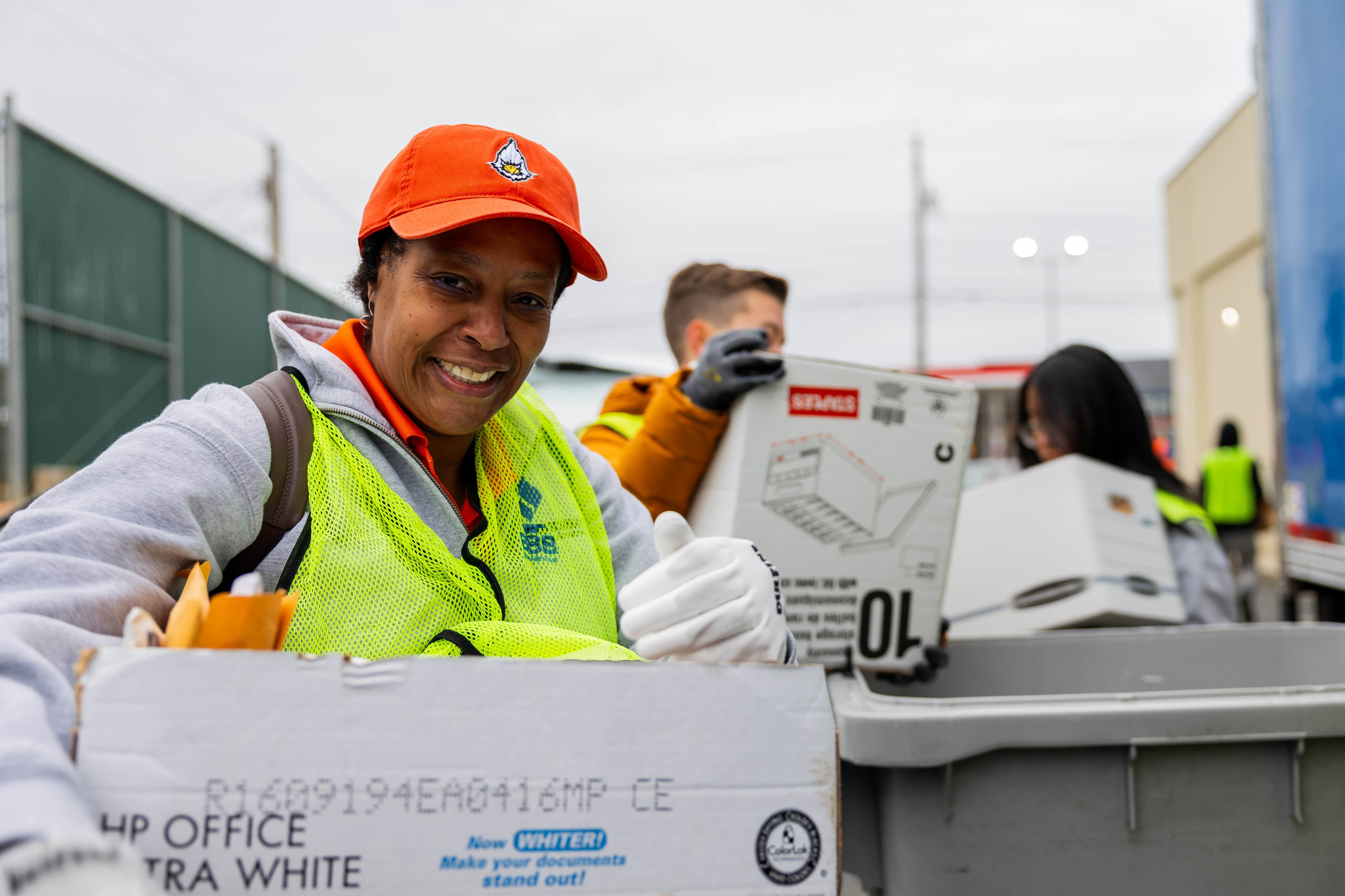 A volunteer in a high-visibility vest and gloves, smiling as she carries a box of documents at Shred Day.